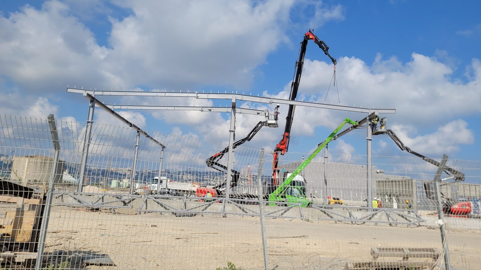 הקמת שלד מבנה פלדה באתר בנייה עם מנוף ובמות הרמה, ברקע נוף חיפה